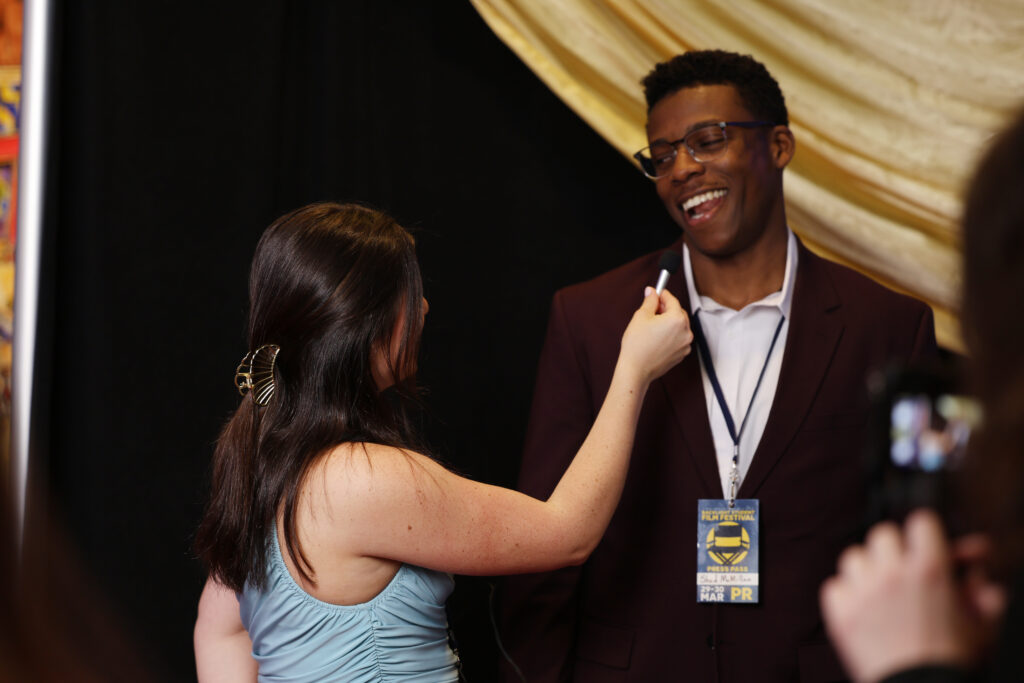 A woman holds a microphone up to a smiling man, Shad McMillan, who wears glasses and a suit with an event badge around his neck. They stand in front of black and gold draped fabric.