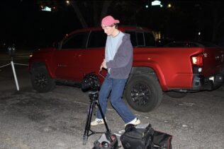 Aidan Landrum, wearing a pink cap, gray hoodie, and blue jeans, sets up a camera on a tripod in a parking lot at night, with a red pickup truck and camera bags nearby.