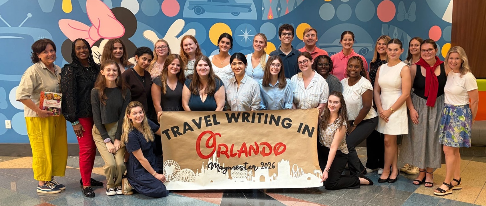 A large group holds up a banner that reads "travel writing in Orlando Maymester 2026."