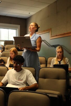 A woman stands up to ask a question in her class, among her peers who are sitting down in a classroom.