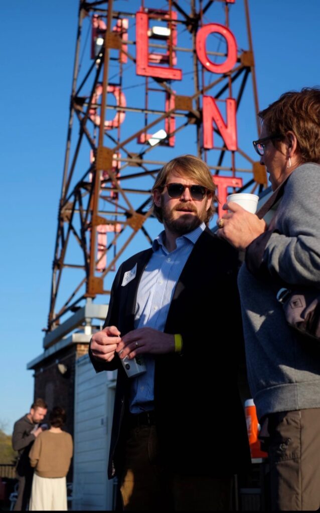 Eric NeSmith, in sunglasses and a blazer, talks with a woman holding a coffee cup in front of a tall metal structure with large red letters. Two other people converse in the background under a clear blue sky.