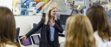 Kim Landrum stands in the graphics lab and points to a computer while students in the foreground look on.