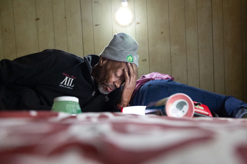 A man wearing a hat lays down and holds his head. There are food containers in the foreground.