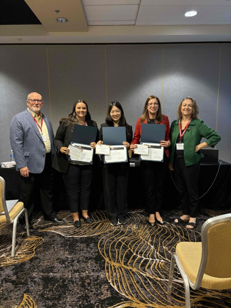 A photo of one man and four women standing in a conference room. Three of the women are holding some type of award.