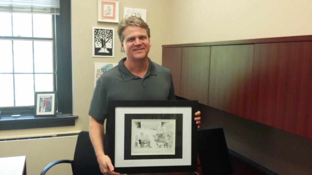 Wilson Lowrey is pictured in an office holding a political cartoon he drew during his time at the Athens Banner-Herald.