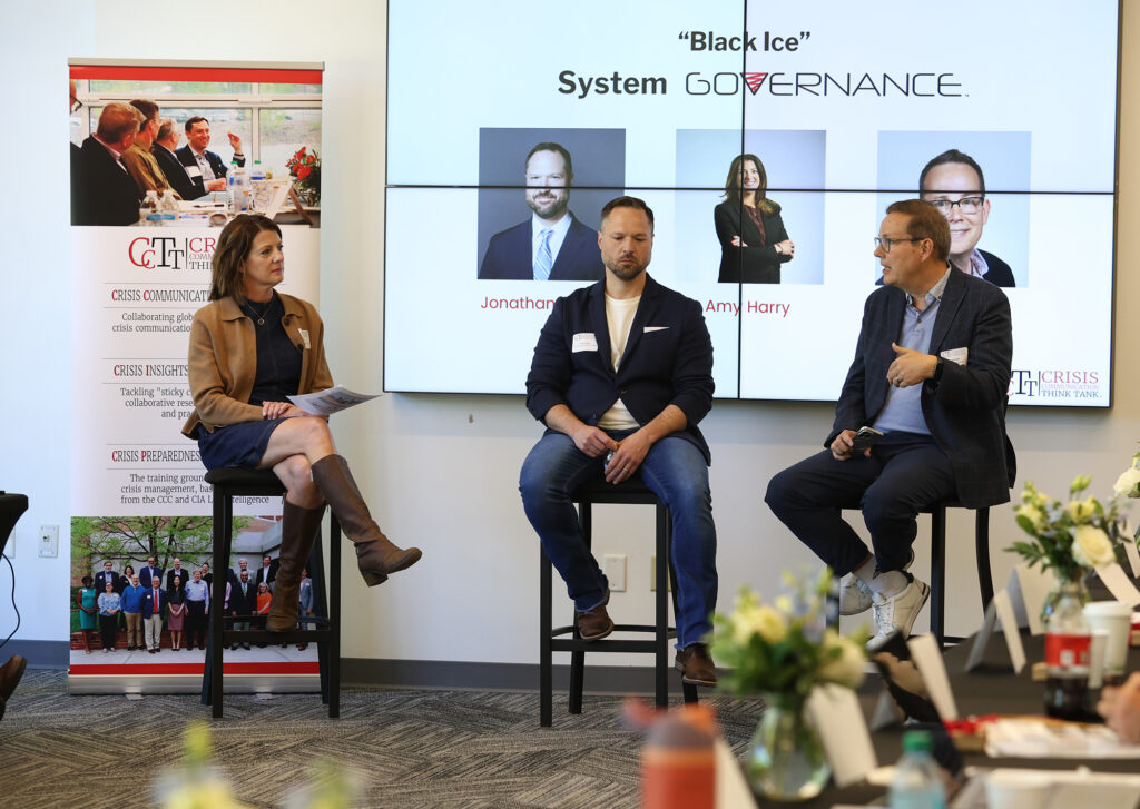 A panel titled "Black Ice: System Governance." Three participants sit on stools. On the left, a woman in a brown cardigan and navy dress holds a microphone. In the center, a man in a navy blazer and white t-shirt looks on. The screen behind them shows headshots.