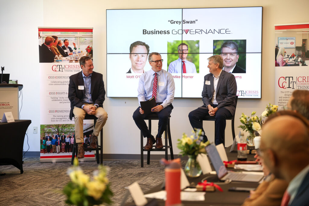 A panel titled "Grey Swan: Business Governance." Three men are seated on stools, laughing together. The man on the left is in a navy blazer and tan khakis; the center man, Mike Pferrer, wears a light blue shirt and red striped tie; the man on the right is in a grey suit. The screen behind them displays their professional headshots.