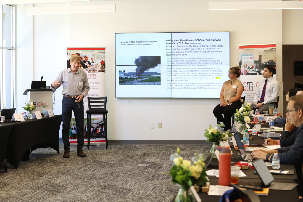 A man in a grey button-down shirt and navy trousers presents at the front of the room, gesturing toward a screen displaying a news report about a UPS plane crash and Boeing. Two other participants, a woman in a tan top and a man in a white shirt and tie, sit to the right observing the presentation.