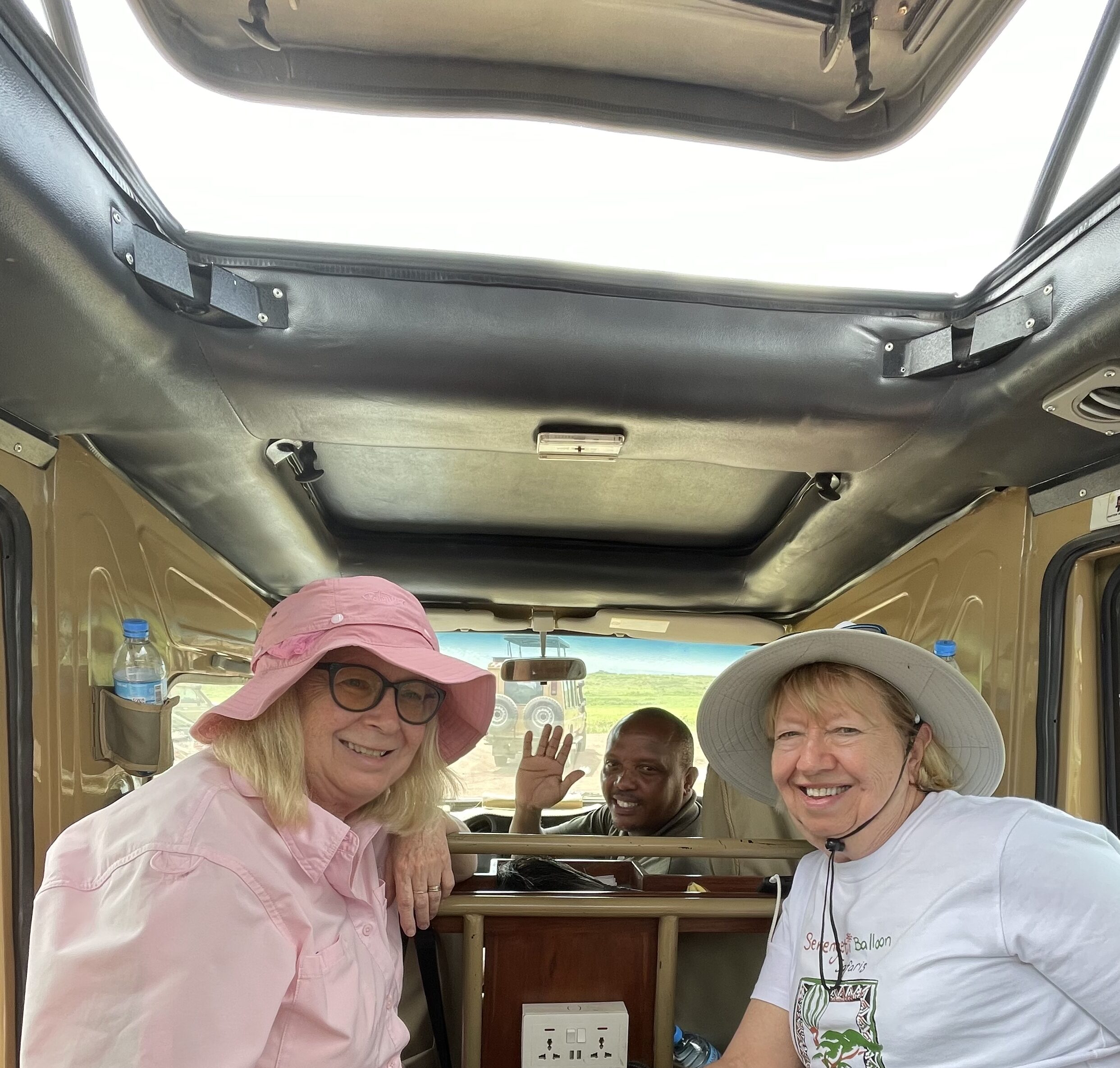 Carolyn Tieger (pictured on the right) travels in a safari vehicle with another passenger and driver waving in the background.