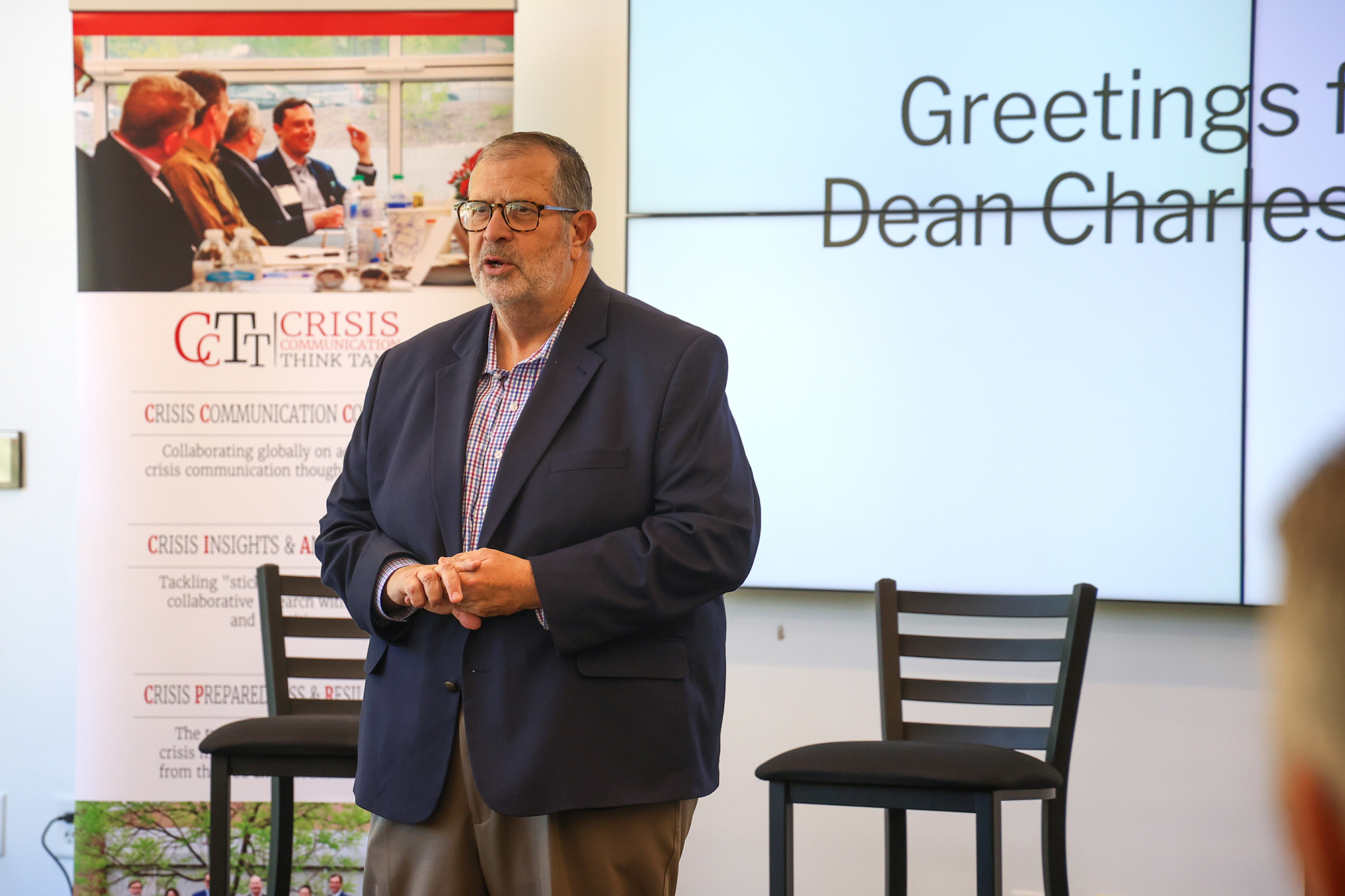 A close-up of Dean Charles Davis speaking in front of a group. He is a man with glasses and a beard, wearing a navy blue blazer over a checkered shirt. The background shows a blurred digital screen with the text "Greetings from Dean Charles..." and a CCTT banner to the left.