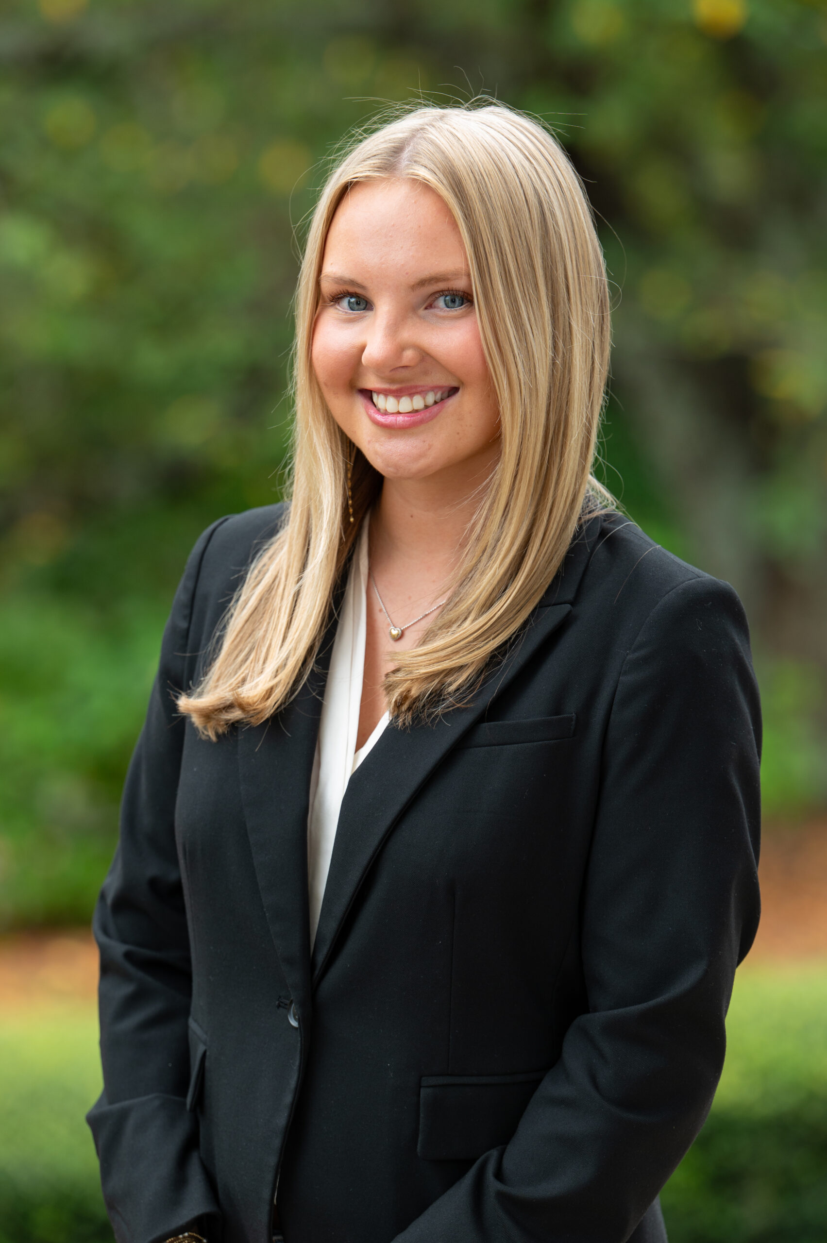 A young woman with long blonde hair wearing a black blazer and white blouse stands outdoors, smiling at the camera with a blurred green background.