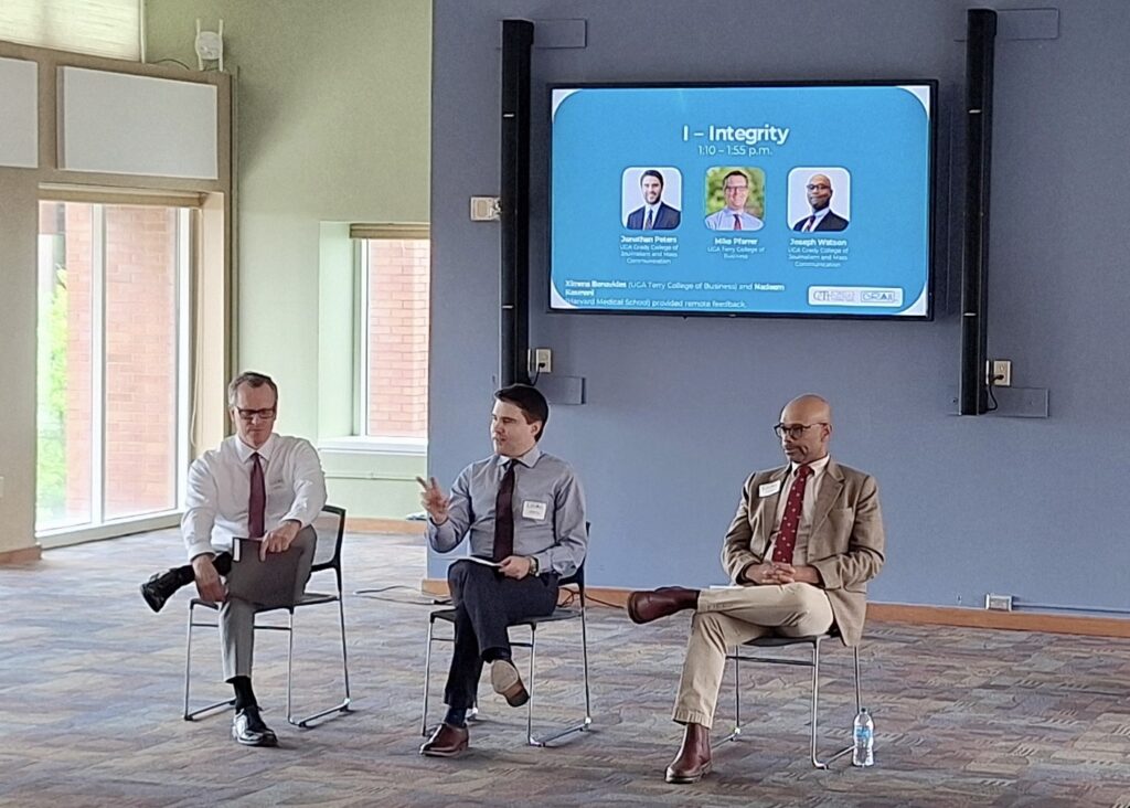 Three men sit and present in front of a digital panel that reads I - Integrity.