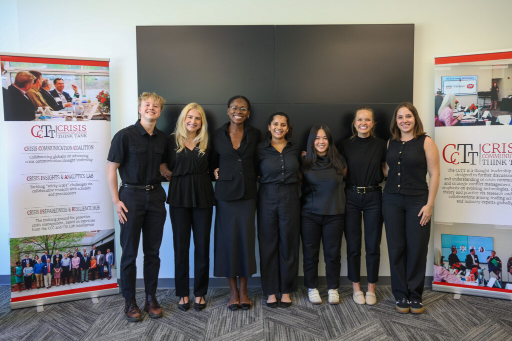 A group of students all dressed in black, interlock arms and pose for a picture. Two CCTT Crisis Communication Think Tank banner are at their sides.