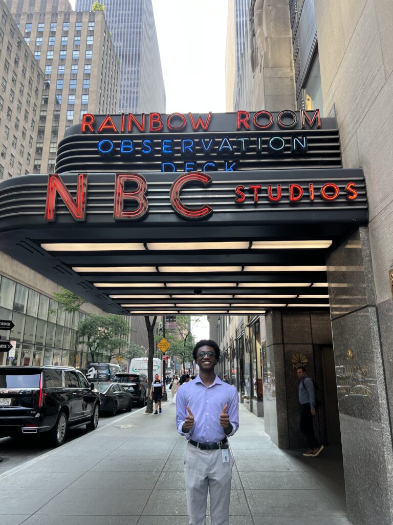 Eniola Badejo stands smiling with two thumbs up under the neon NBC Studios sign at the entrance to the Rainbow Room and Observation Deck in New York City, with tall buildings and pedestrians lining the street.