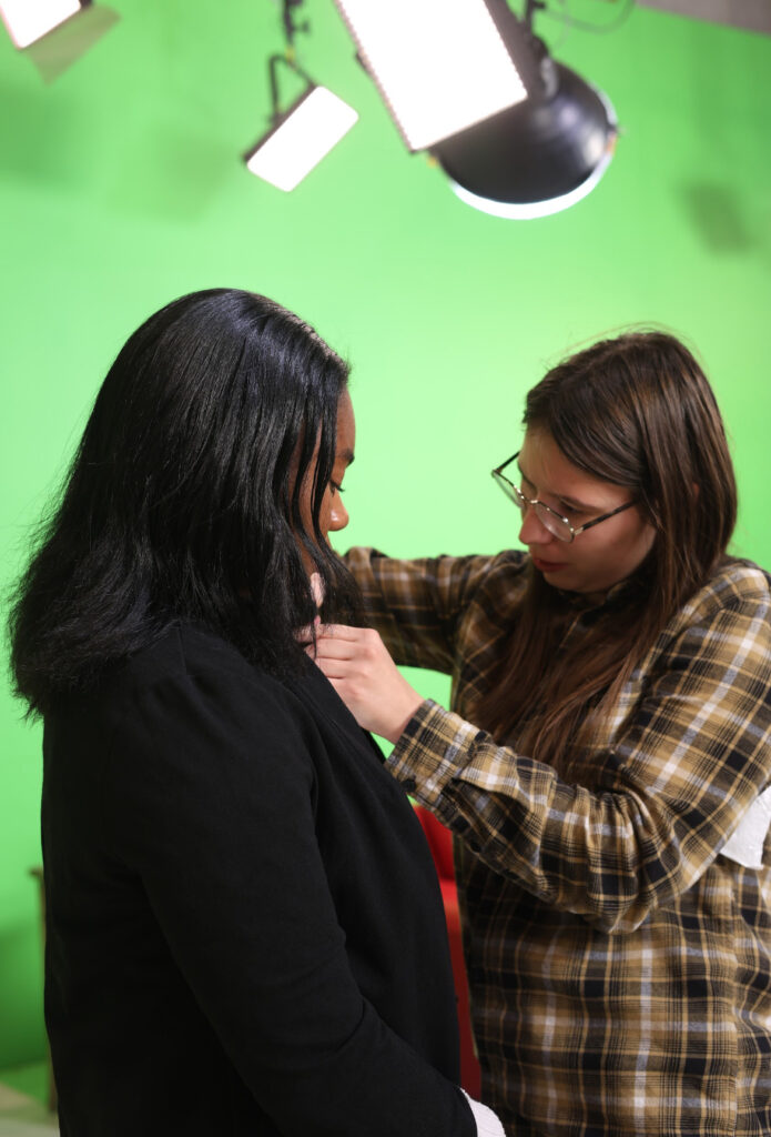 Courtney Craft adjusts a lapel microphone on another woman in front of a green screen, with studio lights visible above them.