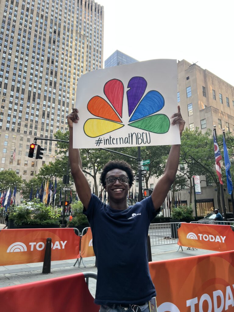 A smiling person, possibly Eniola Badejo, holds up a sign with the colorful NBC peacock logo and hashtag #internalNBCW outside, with tall buildings, trees, and TODAY show banners in the background.