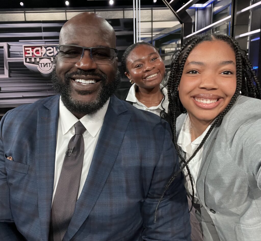 Three people in business attire, including Courtney Craft, smile for a selfie in a TV studio with bright lights and the Inside NBA TNT logo glowing in the background.