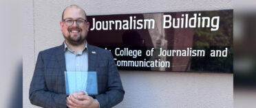 Kyser Lough smiles and holds an award as he poses in front of a sign that reads Journalism Building, College of Journalism and Mass Communication.