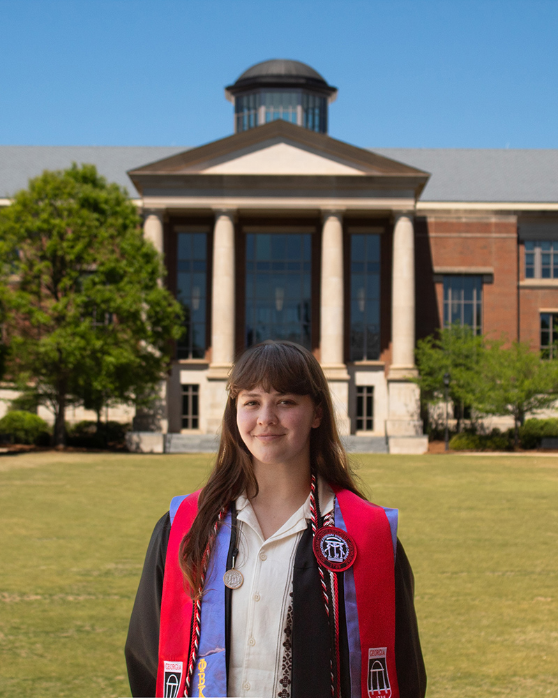 Woman in a graduation gown with honor stoles stands in front of Russell Special Collections Library on UGA campus.
