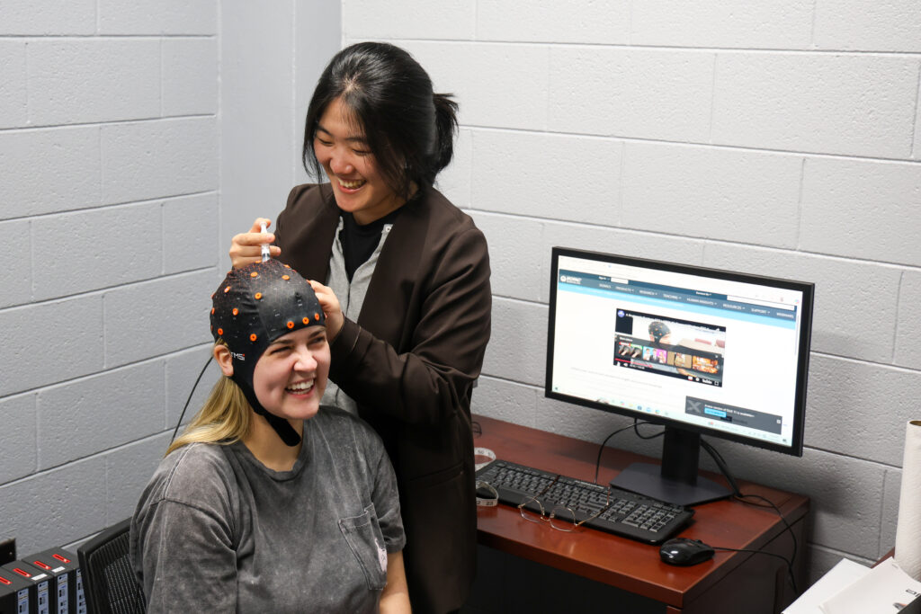 Two women in a neuroscience lab; one smiles, wearing a sensor cap as Brittany Shivers adjusts it. Behind them, a computer displays research websites, highlighting their focus on brain science and innovation.