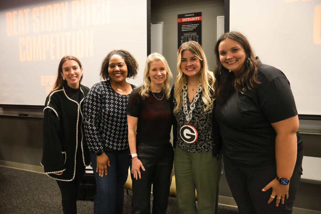 Five women, including Brittany Shivers, stand and smile together indoors in front of a screen displaying “Beat Story Pitch Competition.” The woman in the middle proudly holds a “G” medallion at this lively event.