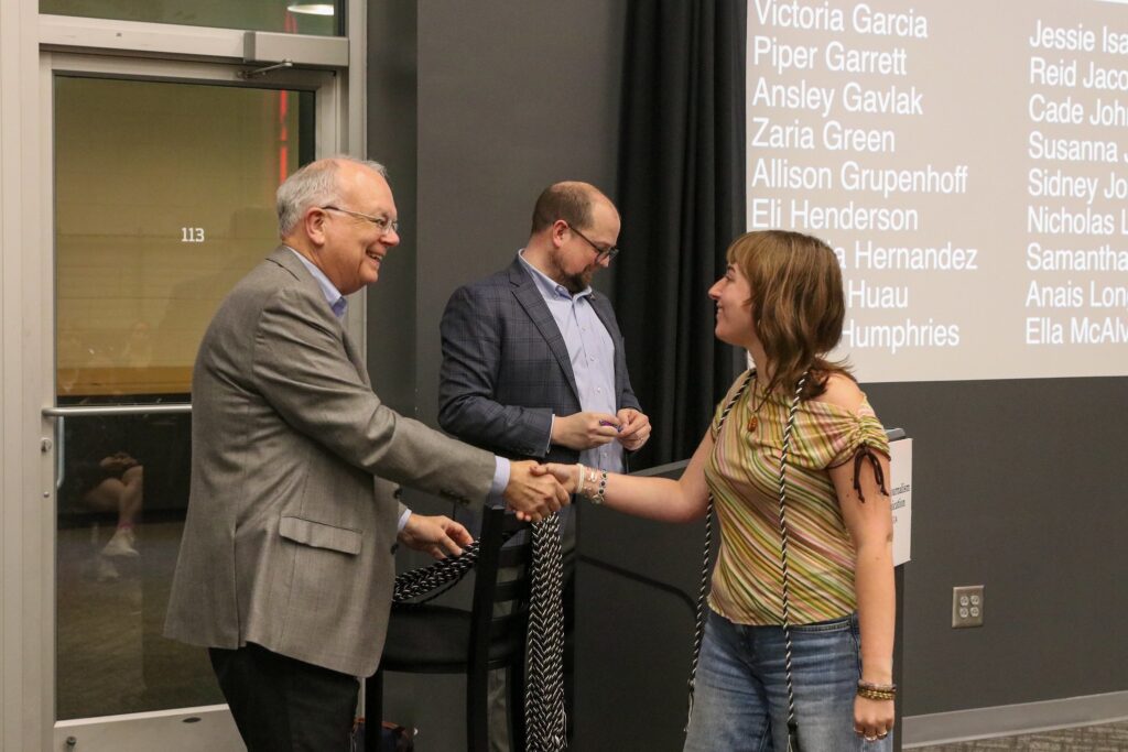 A student shakes hands with a professor. The student has a graduation cord around her neck, and the professor is holding a collection of the remaining graduation cords. A second professor stands in the background behind a podium and in front of a presentation projected on the wall.