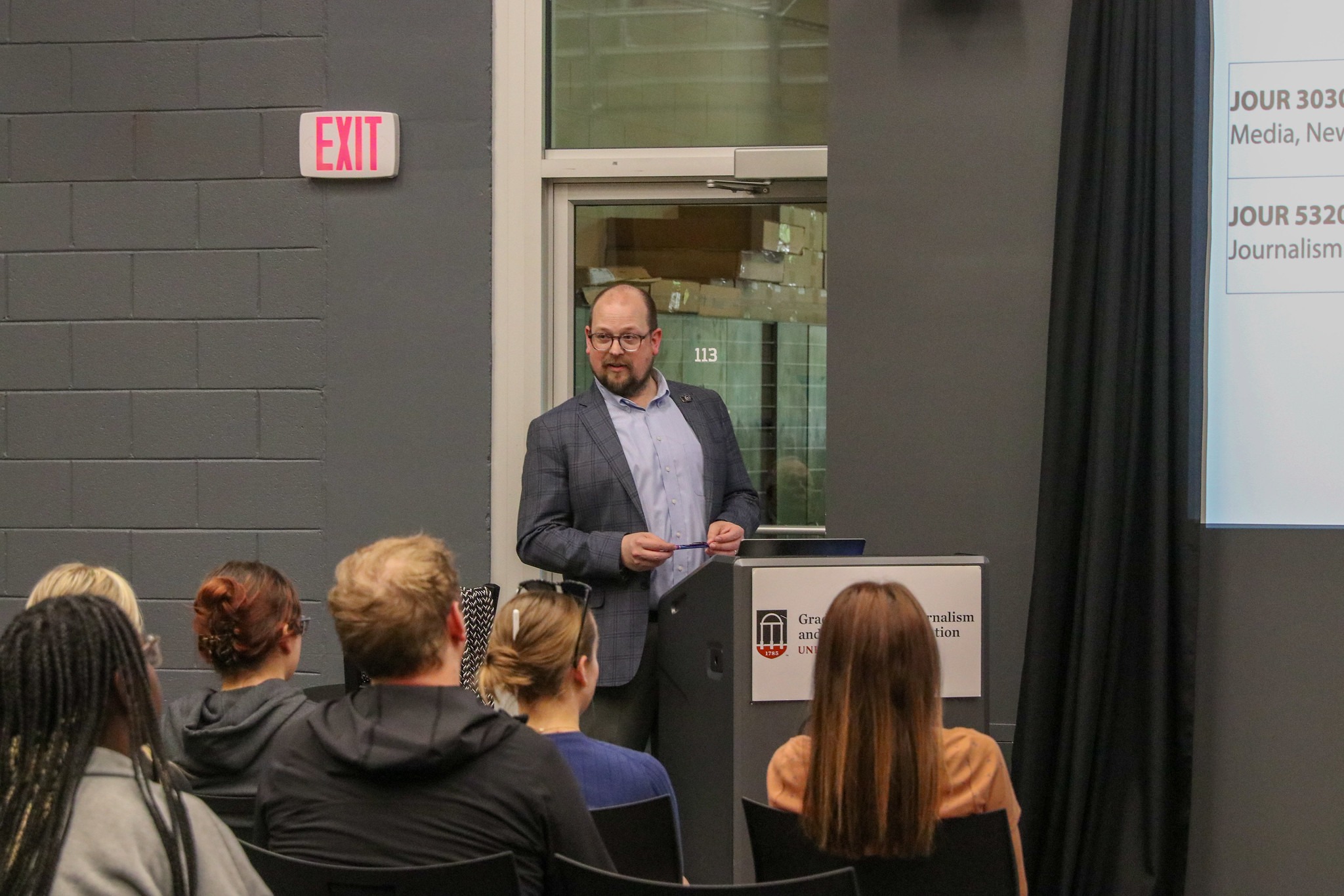 A man stands at a podium in front of a group of students, speaking in front of a presentation projected on the wall behind him.