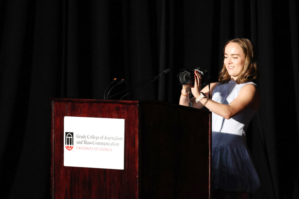A woman standing at a podium holds a camera and takes a picture. She stands behind a lectern with a sign reading Grady College of Journalism and Mass Communication.