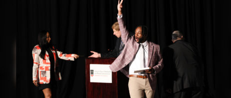 A man gives an YO hand sign for his fraternity Kappa Alpha Psi as he crosses a stage. He is holding a box in his hand and there is a lectern in the background reading Grady College of Journalism.