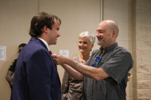 A candid moment where an older man in a patterned shirt adjusts the tie of a younger man in a navy blue suit. An older woman with short grey hair and glasses stands between them, watching with a warm smile.