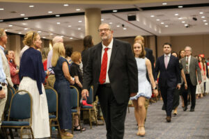 A man in a black suit and red tie leads a procession of graduates and faculty down the center aisle of a large event hall. The audience stands on either side, watching as they walk toward the front.