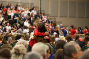 A high-angle, candid shot of a graduate in a red blazer embracing another person in the middle of a large auditorium. The background is filled with a soft-focus audience seated in bleachers and chairs.