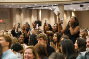 A lively group of young women in an auditorium cheering and holding up their smartphones to take photos or videos. Their expressions are joyful, with some laughing and shouting in celebration.