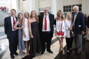A group of eight family members and graduates pose for a multi-generational photo in a modern, bright lobby with marble floors. The group includes two graduates in white dresses and red stoles, surrounded by smiling adults in formal and semi-formal attire.