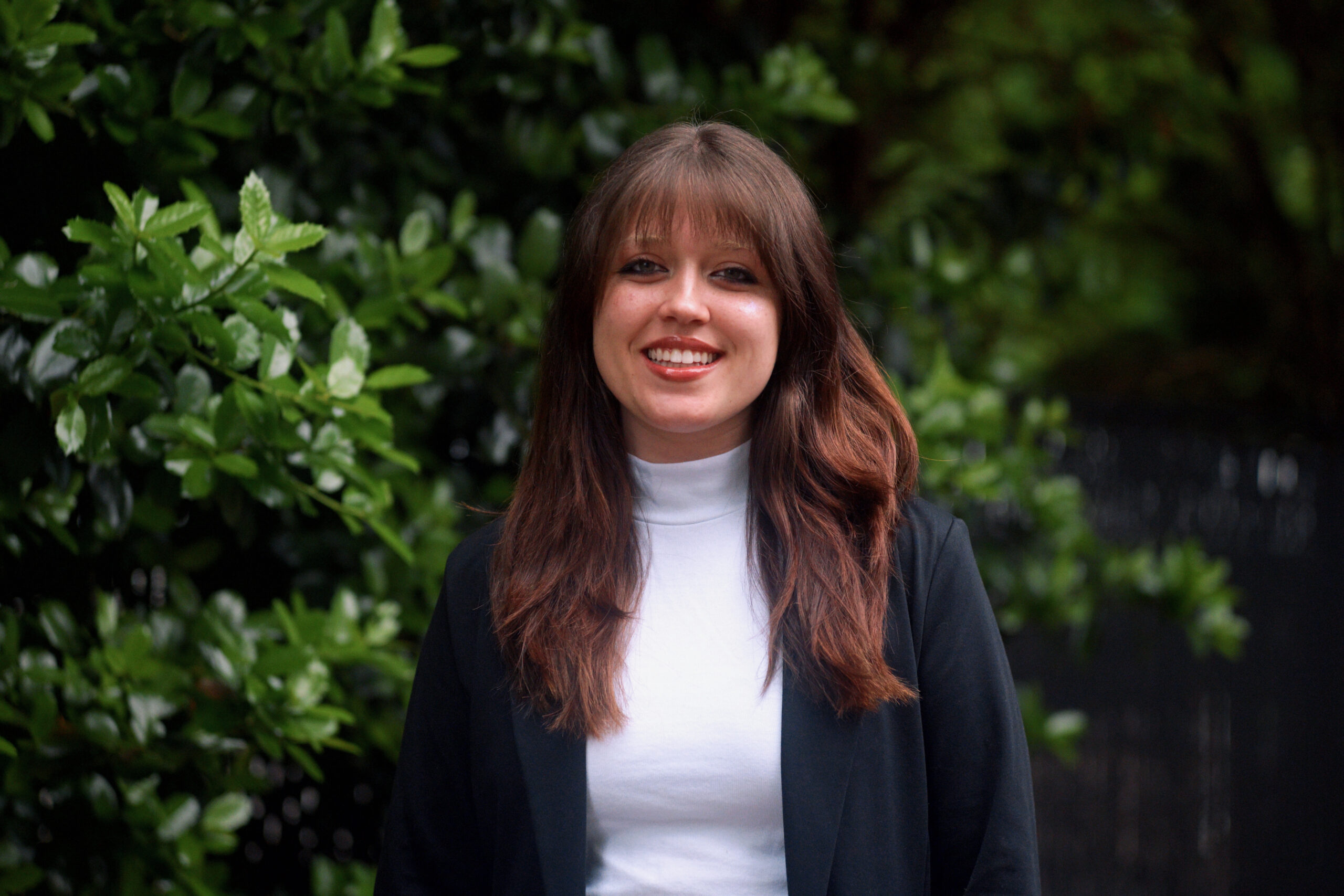 A young woman with long brown hair and bangs smiles at the camera. She is wearing a white turtleneck and a black blazer, standing outdoors in front of green leafy bushes.