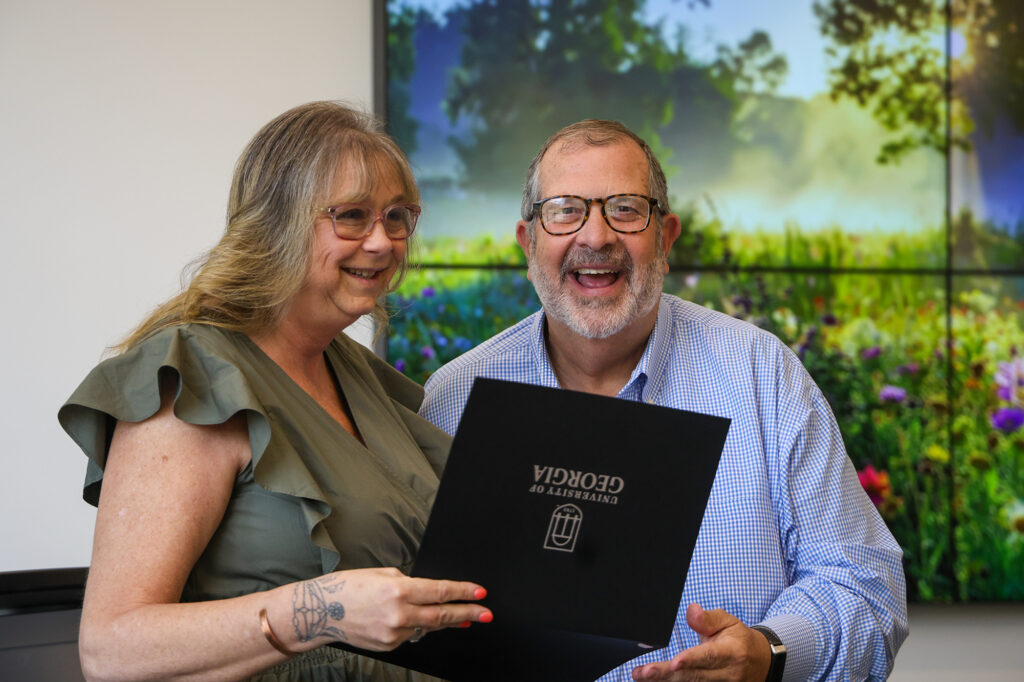 Barbara Bond smiles as she accepts an award in a black folder from Dean Davis. A floral photo is displayed in the background.