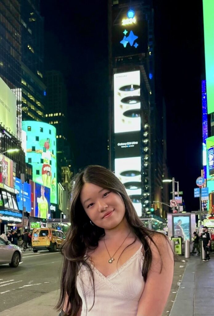 Sophia Kim, a young woman with long dark hair, smiles and poses on a brightly lit street in Times Square at night, colorful billboards and city lights illuminating the background.
