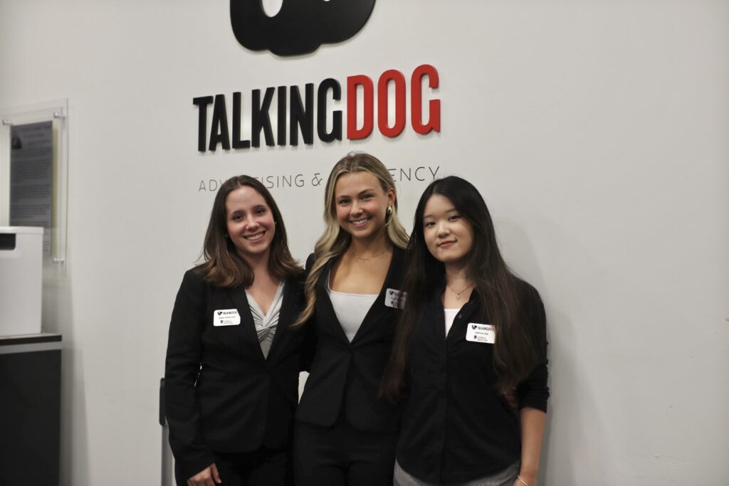 Three women in business attire, including Sophia Kim, stand smiling in front of a wall with the TALKINGDOG Advertising Agency logo. Each wears a name tag and they are posing closely together for the photo.