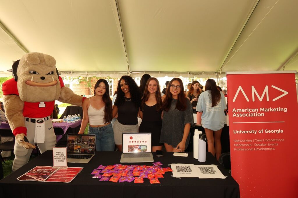 Five people, including Sophia Kim and a mascot in a bulldog costume, stand smiling behind a table with marketing materials and candy. A red American Marketing Association sign for the University of Georgia is displayed on the right.