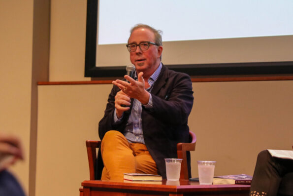 A man sitting in a chair in front of a crowd speaks into a microphone.
