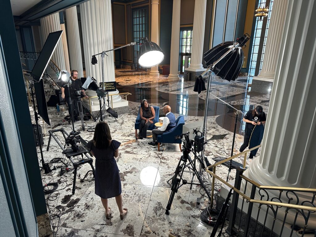 An image of a film set full of equipment with one woman standing and two people seated.
