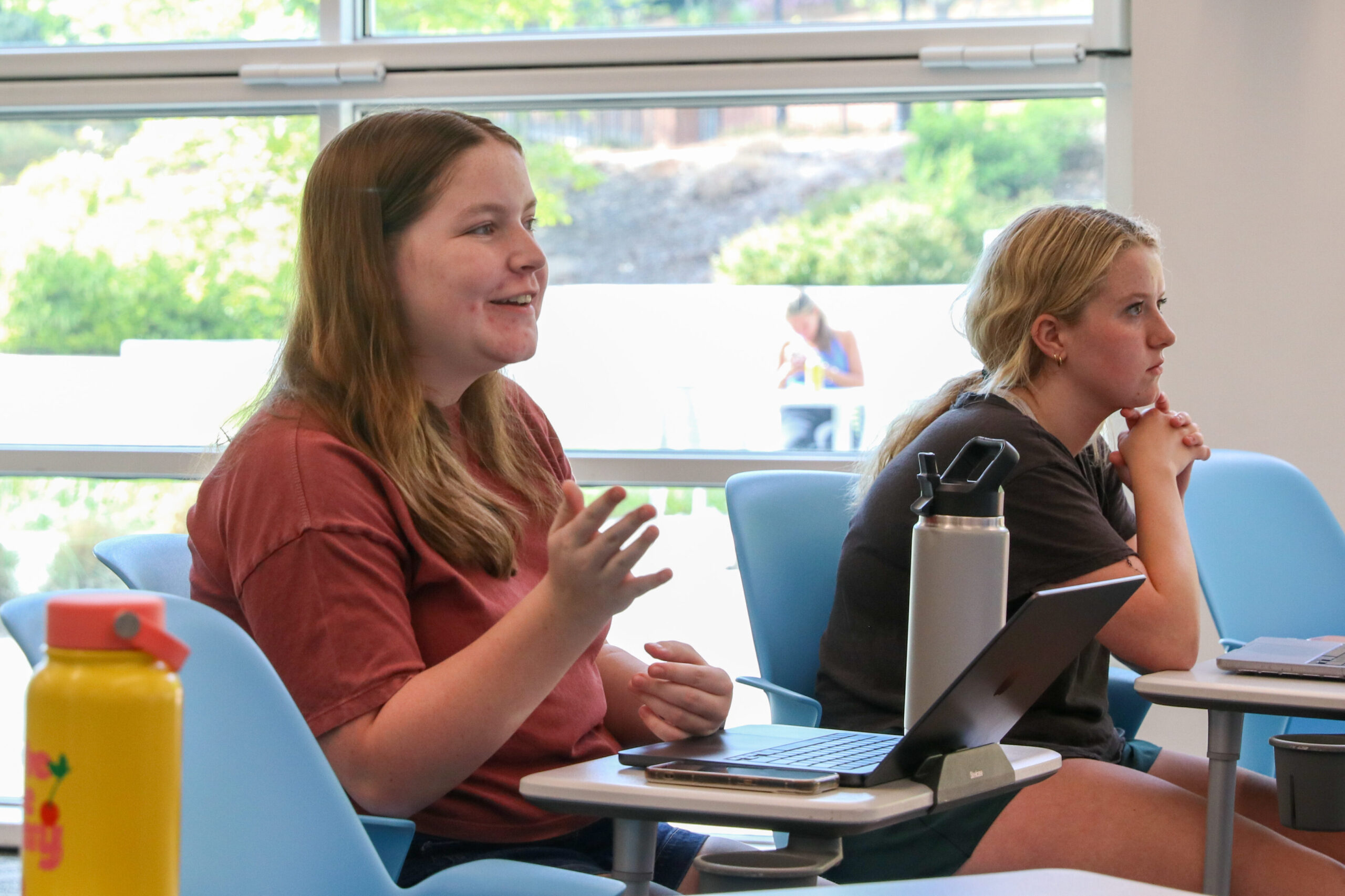 A medium shot of two young women sitting in a sunlit classroom. The woman in the foreground has long light-brown hair and wears a reddish-pink t-shirt; she is smiling and gesturing with her hands as if speaking or asking a question. Next to her, a blonde woman in a dark grey t-shirt listens intently with her hands clasped near her face. Both are seated in light blue chairs with attached swivel desks holding laptops. Large windows in the background look out onto a green, leafy landscape, and a yellow water bottle is visible in the lower left corner.