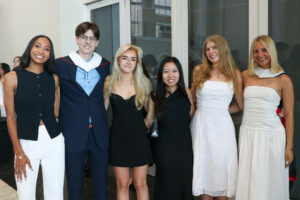 Six young adults stand in a row posing for a group photo in a modern hall. They are dressed in semi-formal attire, including black and white dresses and a blue suit. Two of the individuals wear white academic hoods over their shoulders.