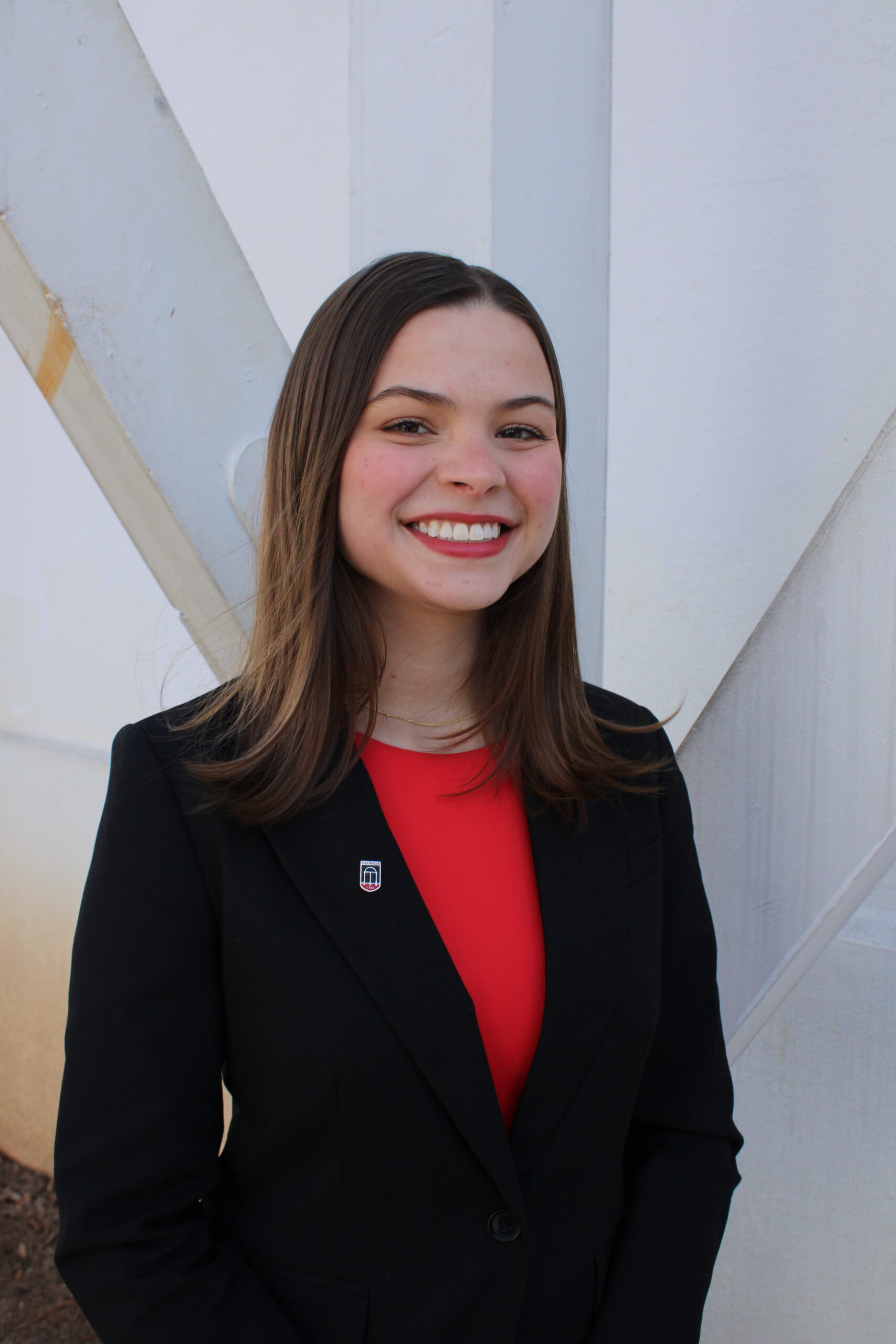 A young woman with straight brown hair, wearing a black blazer over a red top, stands smiling in front of a white wall with intersecting beams.