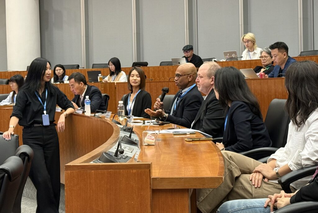 A man speaks sitting down with a microphone infant of a lecture hall. 
