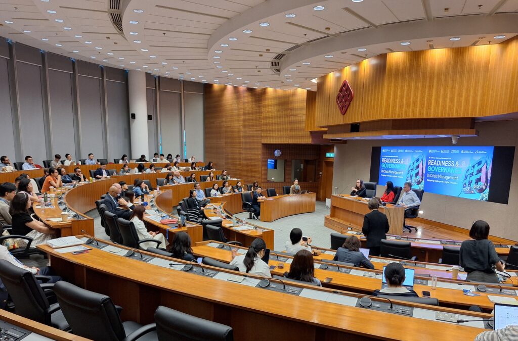 A wide-angle shot of a lecture hall full of people listening to a presentation with three speakers in the front. 