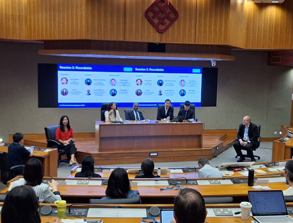 A group of guest speakers sit in the front of a lecture hall surrounded by listeners at their desks. 