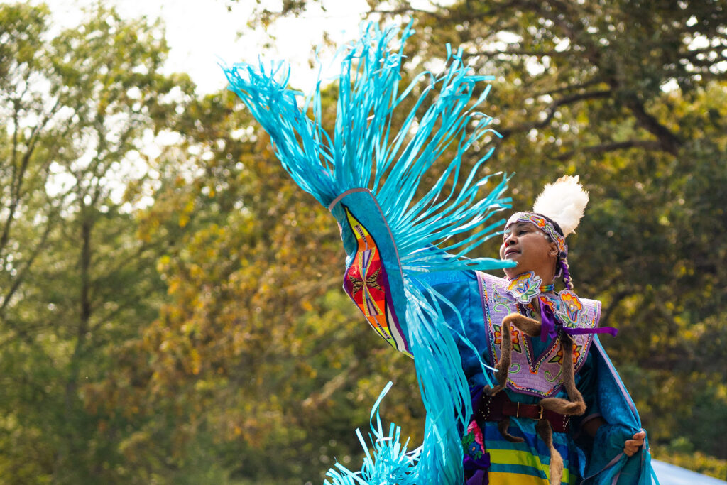 A Native American woman in ceremonial dress waves a blue shawl in a dramatic flair during a Powwow.