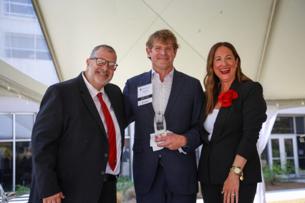 Two men and a woman stand together under a tent. The man in the middle holds a trophy.