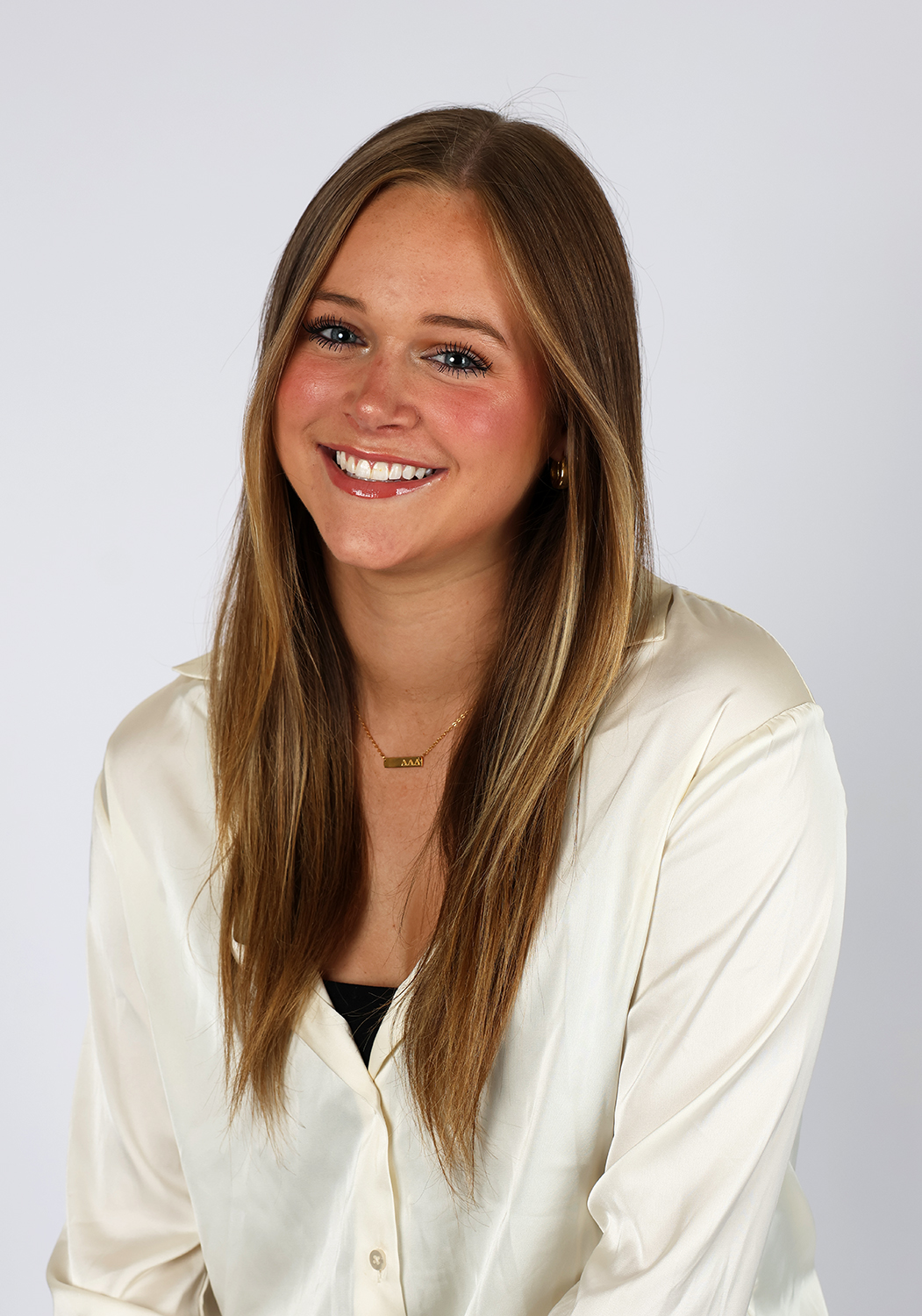 A young woman with long, straight light brown hair smiles at the camera. She is wearing a cream-colored blouse and a gold necklace, seated in front of a plain white background.